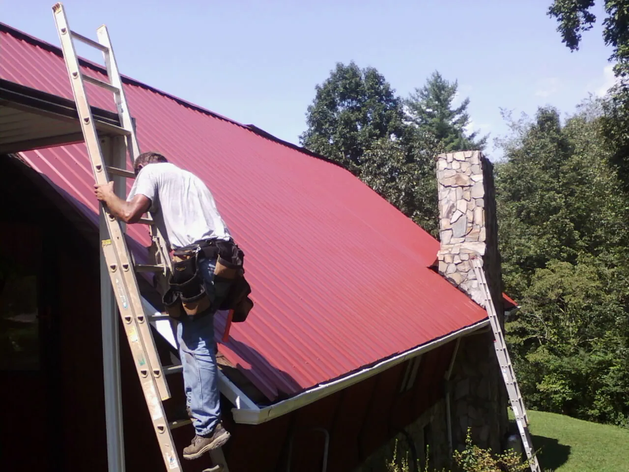 Roofer on a ladder beside a red metal roof and stone chimney — Soffit & Fascia Repair by Roofing Brothers in New River